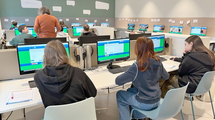 Rückenansicht von einem Raum mit Tischen, auf denen Computer stehen. An jedem Computer sitzt ein Teenager. Neben einem Teenager steht eine PErson