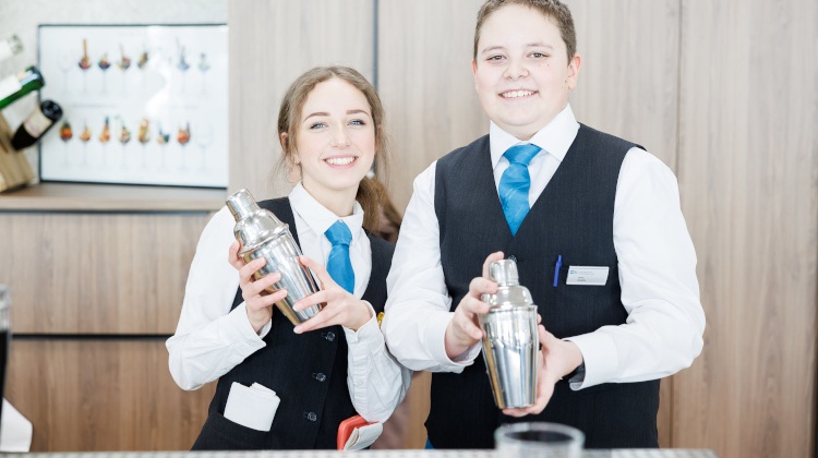 Zwei jugendliche Personen in Arbeitskleidung blicken freudig in die Kamera, während sie Cocktailshaker in Händen halten.