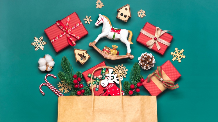 Top-Shot eines braunen, liegenden Sackerls auf einem grünen Hintergrund. Aus dem Sackerl ragen Tannenzweige, Zuckerstangen, Geschenke, Dekosterne. Darüber liegen ein kleines Schaukelpferd, Geschenke, ein Tannenzapfen sowie weitere verpackte Geschenke