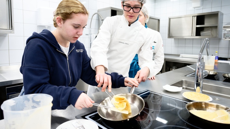 In einer Gastronomieküche stehen neben einer Herdplatte. Eine jugendliche Person sowie eine Person mit Haube, Brille und Schürze. Auf der Herdplatte steht eine Pfanne. Die jugendliche Person und die andere Person kochen darin gemeinsam ein Omlette