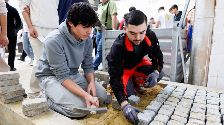In einem kleinen Bereich, der von niedrigen Betonklötzen begrenzt wird, hocken nebeneinander zwei Personen. Die rechte Person legt kleine Pflastersteine auf den Bereich, der mit Sand ausgelegt ist. Die linke Person hält eine Spitzhacke