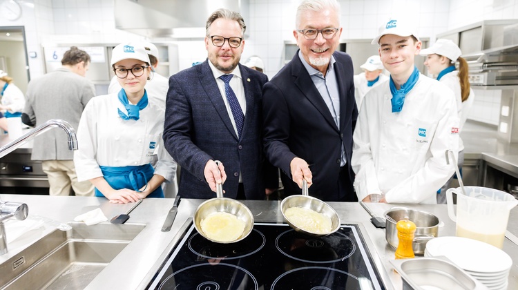 Porträt von zwei Personen in dunklen Sakkos und mit Bart, die hinter einem Ceranfeld stehen und jeweils eine Pfanne halten. Neben ihnen stehen zwei jugendliche Personen in Kochkleidung. Im Hintergrund sind weitere Personen