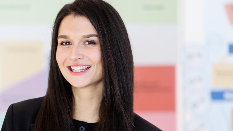 Porträt einer lächelnden Person mit offenen, dunklen Haaren sowie dunklem Oberteil vor hellem Hintergrund