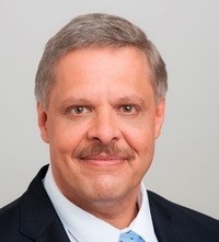 Porträt einer lächelnden Person mit grauen Haaren, Schnurrbart, Hemd, hellblauer Krawatte sowie Sakko vor hellem Hintergrund