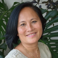 Portrait von Colette Fischer, vor einer Zimmerpflanze stehend und lächelnd