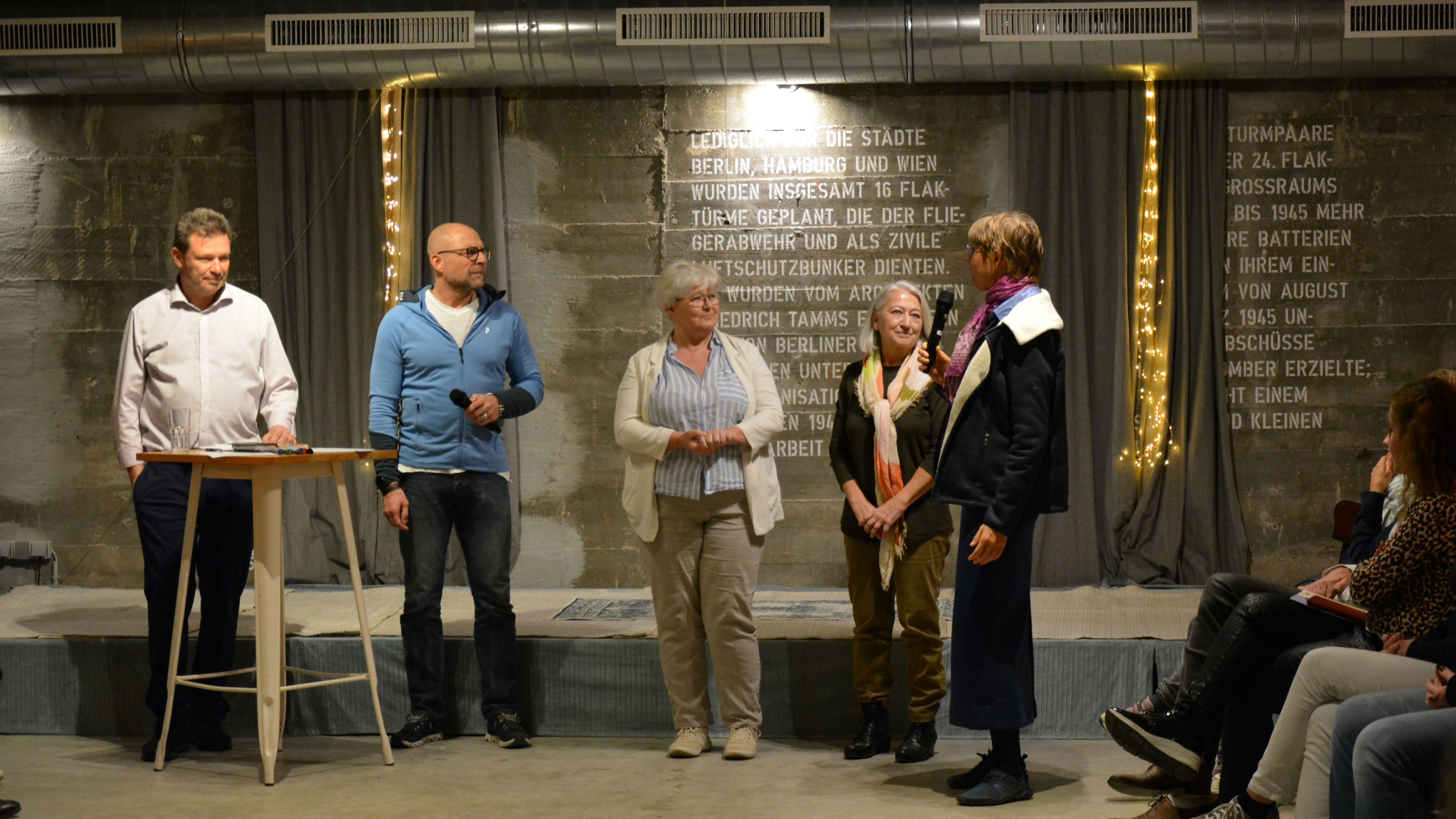 Personen stehen vor einer kleinen Bühne und sprechen miteinander, das Publikum hört zu.