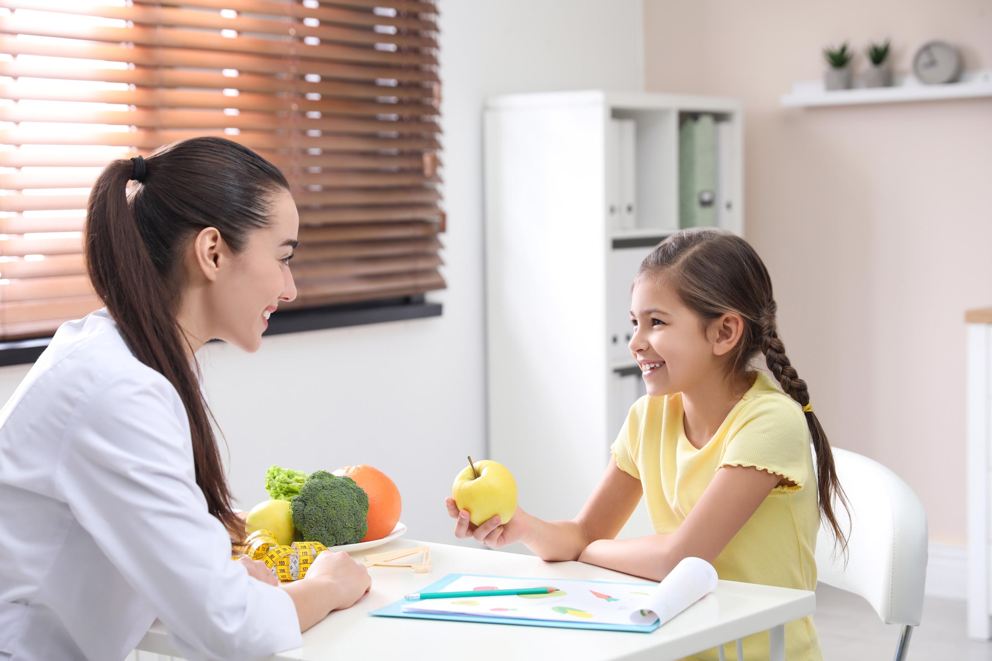 Lächelndes Kind sitzt an Tisch erwachsener, lächelnder Person in weißem Kittel gegenüber und hält in einer Hand gelben Apfel, am Tisch Clipboard mit Stift, Maßband und Schale mit Gemüse und Obst platziert