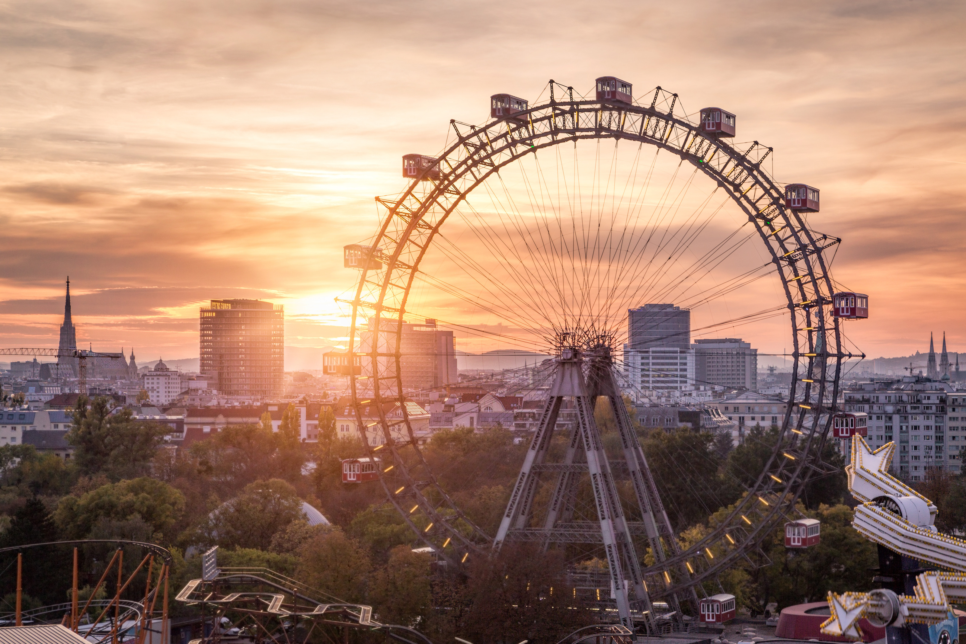 Großes Riesenrad im Wiener Prater mit roten Gondeln im Licht des Sonnenuntergangs, ringsum Baumkronen und im Hintergrund Gebäude der Stadt