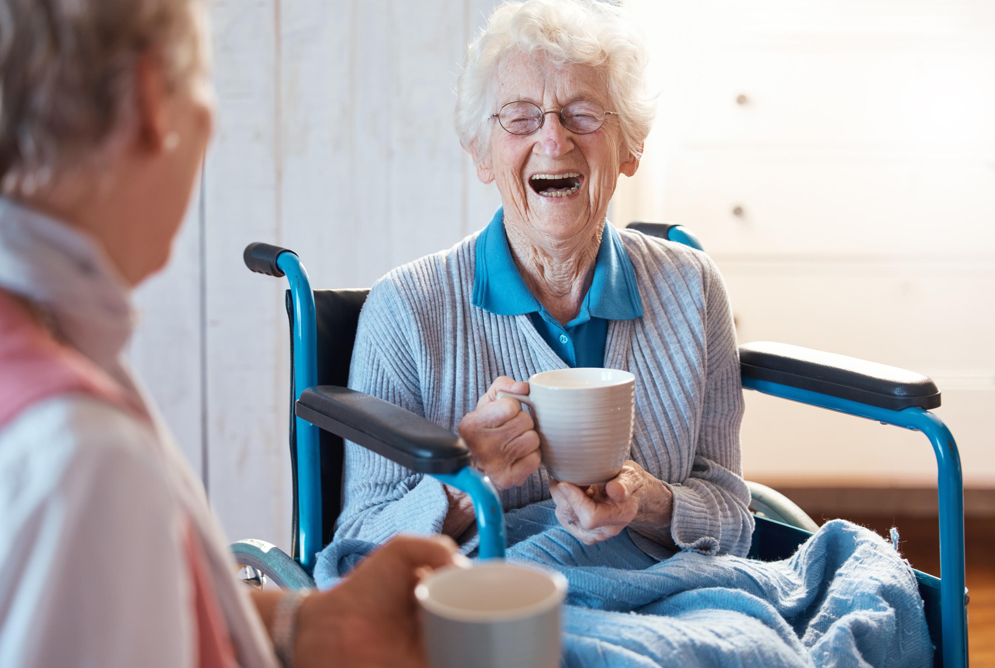 Lachende ältere Person mit Brillen sitzt in Rollstuhl und hält Tasse in der Hand, im Vordergrund verschwommen weitere Person mit Tasse