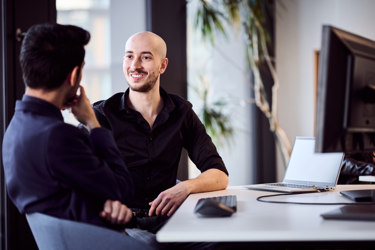 Zwei Personen unterhalten sich, eine davon ist von vorne, die andere von hinten zu sehen. Sie sitzen an einem weißen Tisch, auf dem auch eine Tastatur, eine Computermaus, ein Laptop und ein Monitor platziert sind,