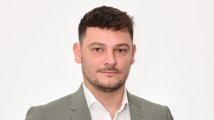 Porträt einer Person mit kurzen Haaren, Bart, hellem Oberteil und grauem Sakko, die vor einem hellen Hintergrund steht