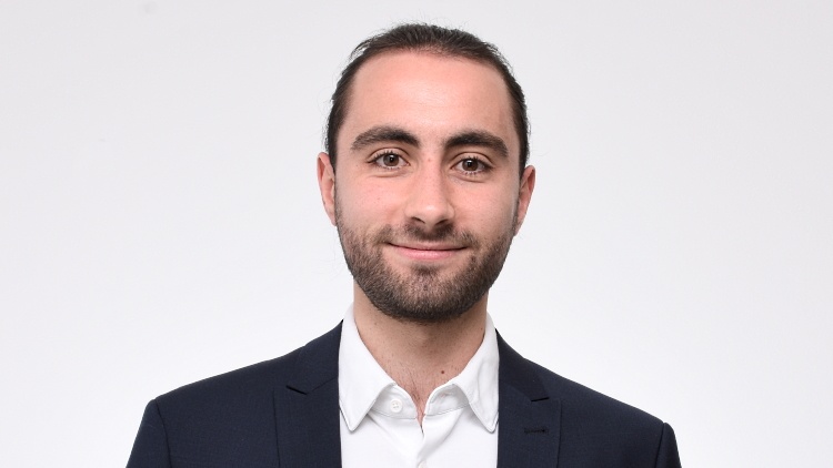 Porträt einer Person mit dunklem, kurzem Haar, Bart, weißem Hemd und dunklem Sakko vor einer weißen Wand